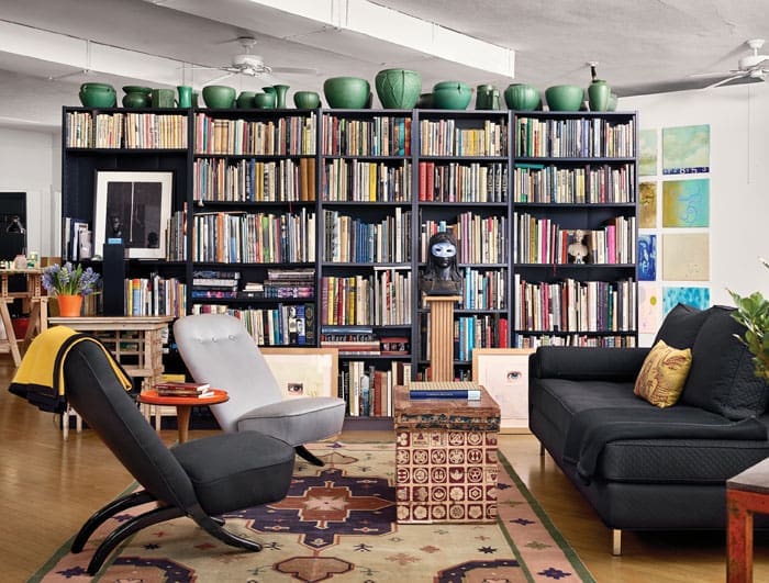 A seating area anchors the space and a massive double-sided bookcase. Daybed, surrounded by Scandinavian mid-century prototype chairs never put in production, alongside Deco bench. Japanese 1920s tea box serves as table. A cotton dhurrie warms the floor. Collection of Arts and Crafts pottery atop the shelves. Bust of Virgin Mary, circa 1940s, with a Palazzolo-made mask for a Magritte table at the annual Inprint Poets and Writers Ball.  
