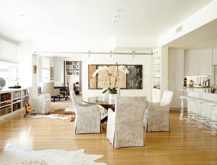 In the dining area, a dining table from Scott + Cooner is attended by chairs upholstered in Peacock Alley linen fabric. The vintage Lucite stools were found at an antique store in Dallas and reupholstered in white ostrich. A custom sliding wall acts as a room divider and backdrop for a large painting by Santa Fe artist Marcia Myers. The floors are white pine.