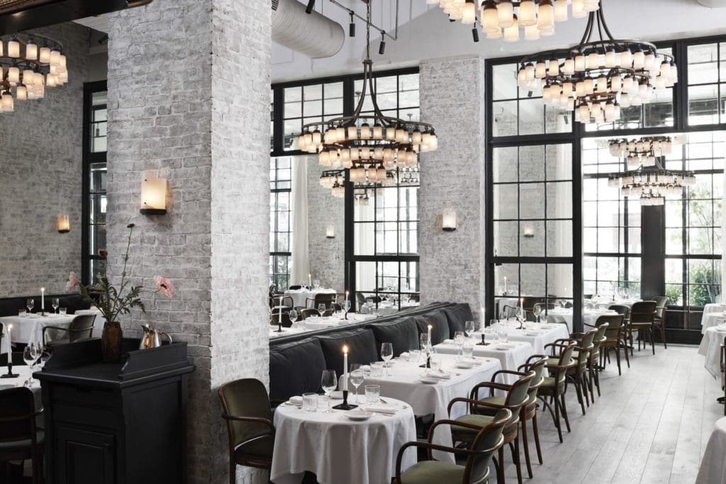 The dining room at Le Coucou features high-exposed concrete ceilings and custom pewtered-steel chandeliers with hand-blown glass shades that mimic candlelight. 