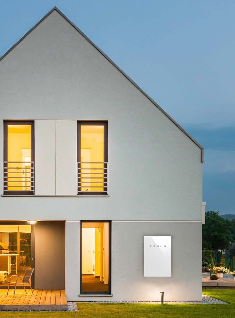 A Tesla Powerwall 2 home battery, shown on an exterior house wall, stores energy from solar panels.