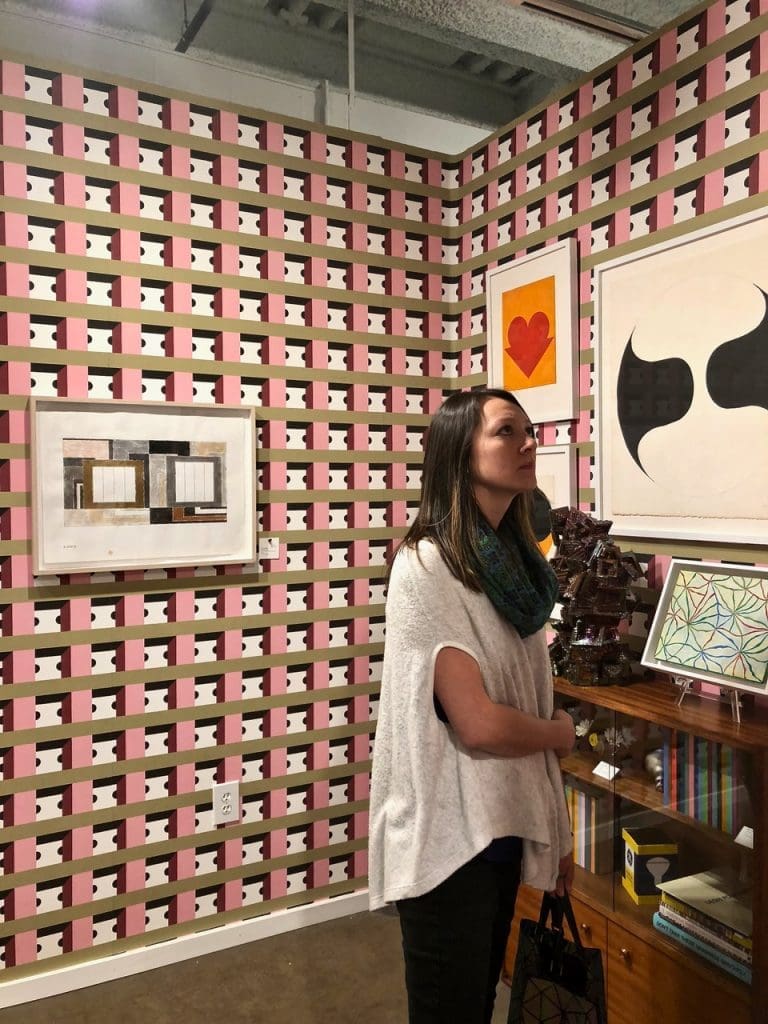 Dr. Cindy Lisica checks out McClain Gallery, including 
wallpaper by artists Nathalie Du Pasquier and George Sowden, from ZigZagZurich, setting a stage for works by  
Peter Halley, Leon Polk Smith, Julia Kunin, Mara Held, Rob Reasoner, and Jonathan Seliger.  
Multiple pieces on background wall (right, in the white frames) by Leon Polk Smith
Shelf: sculpture by Julia Kunin, painting on easel by Mara Held, line paintings inside shelf by Rob Reasoner. Light bulb box sculpture by Jonathan Seliger.