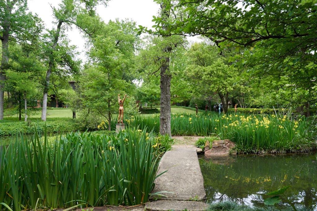 An island in the pond holds a sculpture by Deborah Ballard. The concrete bridge was designed by Donald Vogel. Yellow Louisianna irises growing in the water. (Photo by Pär Bengtsson / art direction by Michelle Aviña)

