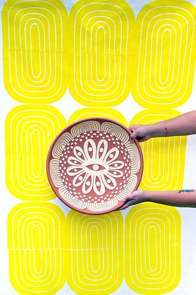 Gabo Martinez holding “Twelve Eyes Platter," 2023. Background: “Yellow Bloom Print,” 2021. On view at Houston Center for Contemporary Craft (Photo courtesy of the artist)