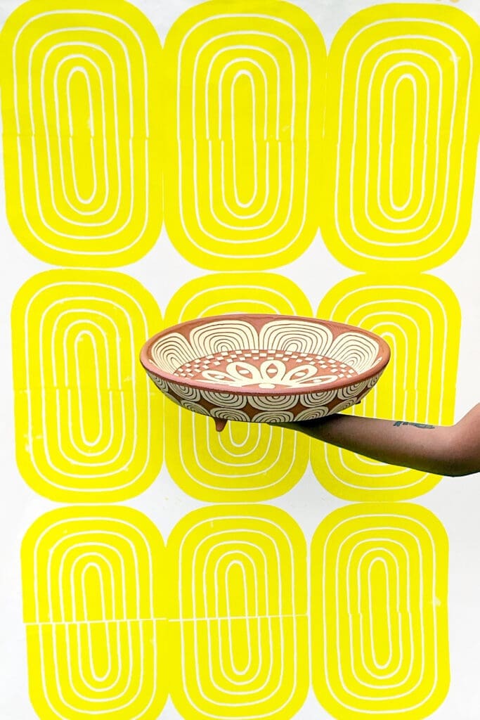 Gabo Martinez holding “Twelve Eyes Platter," 2023. Background: “Yellow Bloom Print,” 2021. On view at Houston Center for Contemporary Craft (Photo courtesy of the artist)
