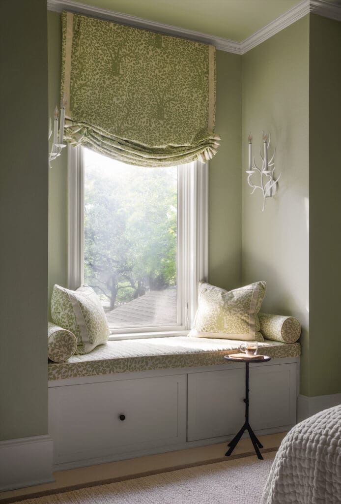 The windows and cushions of this charming nook in a guest bedroom are covered in Quadrille Arbre de Matisse green linen fabric. Walls painted in Benjamin Moore Soft Fern. Global Views table. Visual Comfort sconces. (Photo by Emily Minton Redfield)