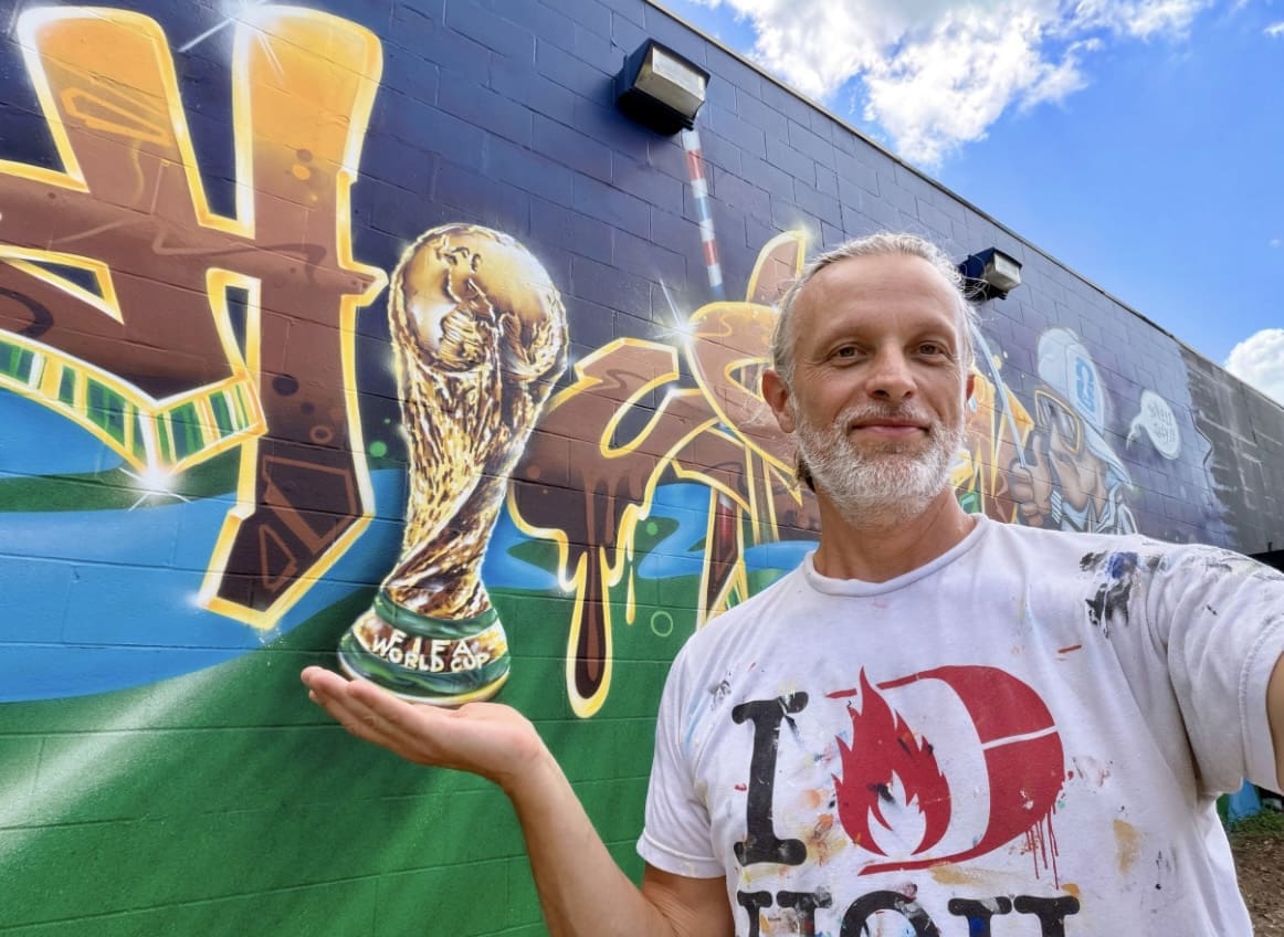 Artist Sebastien "Mr. D 1987" Boileau stands near his mural, ""The Bayou-tiful Game," 2026. (Photo by Eyeful Art)